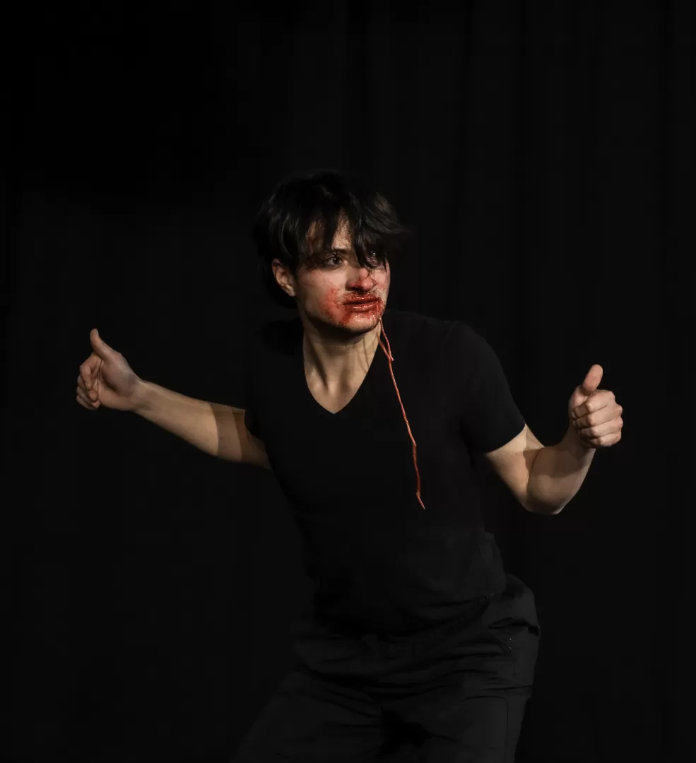 Ein schwarz gekleideter Mann schaut in der Haltung eines Vogels nach rechts. Im Mund hat er rote Schnüre und sein Gesicht ist von einer roten Flüssigkeit verschmiert.