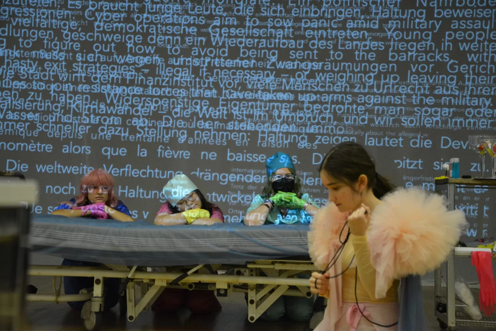 Probenfoto für "Schweig still, mein Stein": Im Vordergrund kniet die Tänzerin und blickt auf den Boden. Die drei Sängerinnen knien hinter einem Krankenhausbett und schauen sie an. Dahinter befindet sich eine Leinwand, auf die eine große Menge Text projiziert ist.