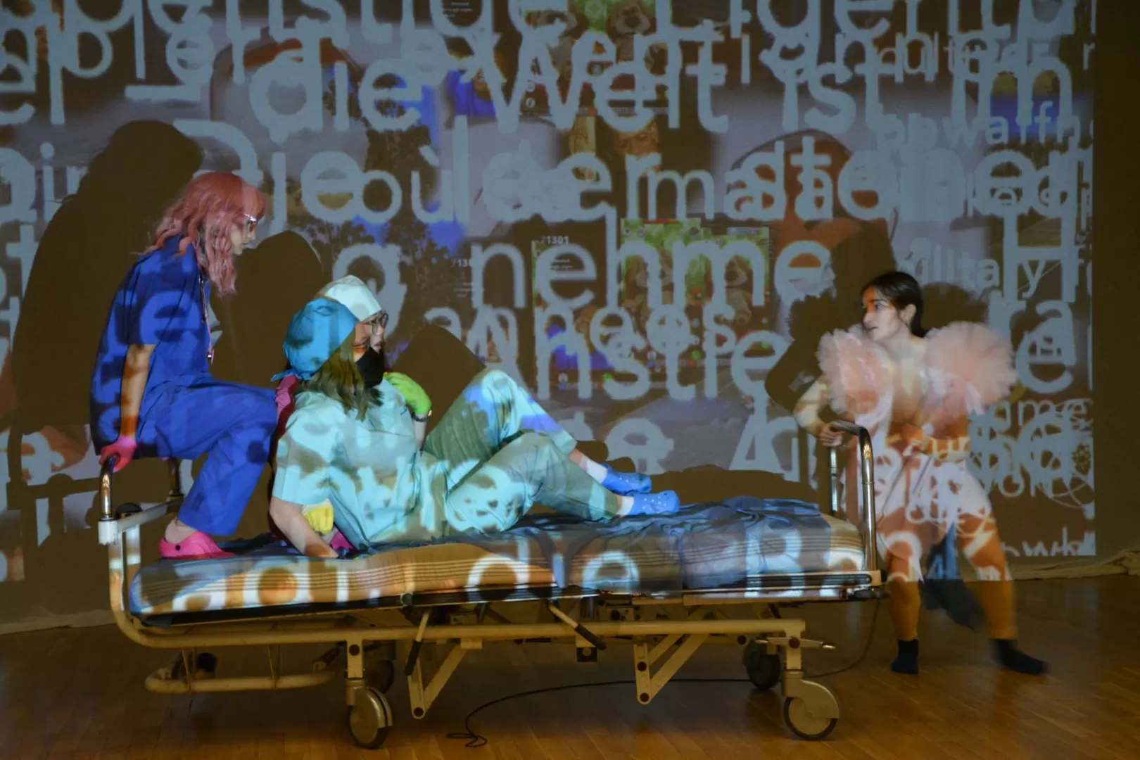 Probenfoto: Drei Darstellerinnen sitzen im Krankenbett, welches von einer vierten gezogen wird. Sie und die Leinwand im Hintergrund werden von einer Projektion mit Text angeleuchtet.