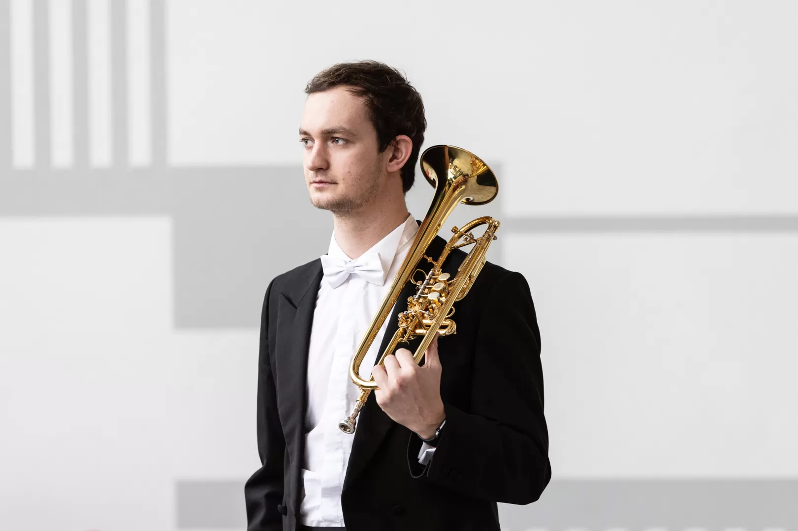 Portrait von Steffen Hillinger. Er trägt einen schwarzen Anzug mit einem weißen Hemd und stützt seine Trompete auf seiner Schulter ab. Er blickt mit ernstem Ausdruck nach rechts.
