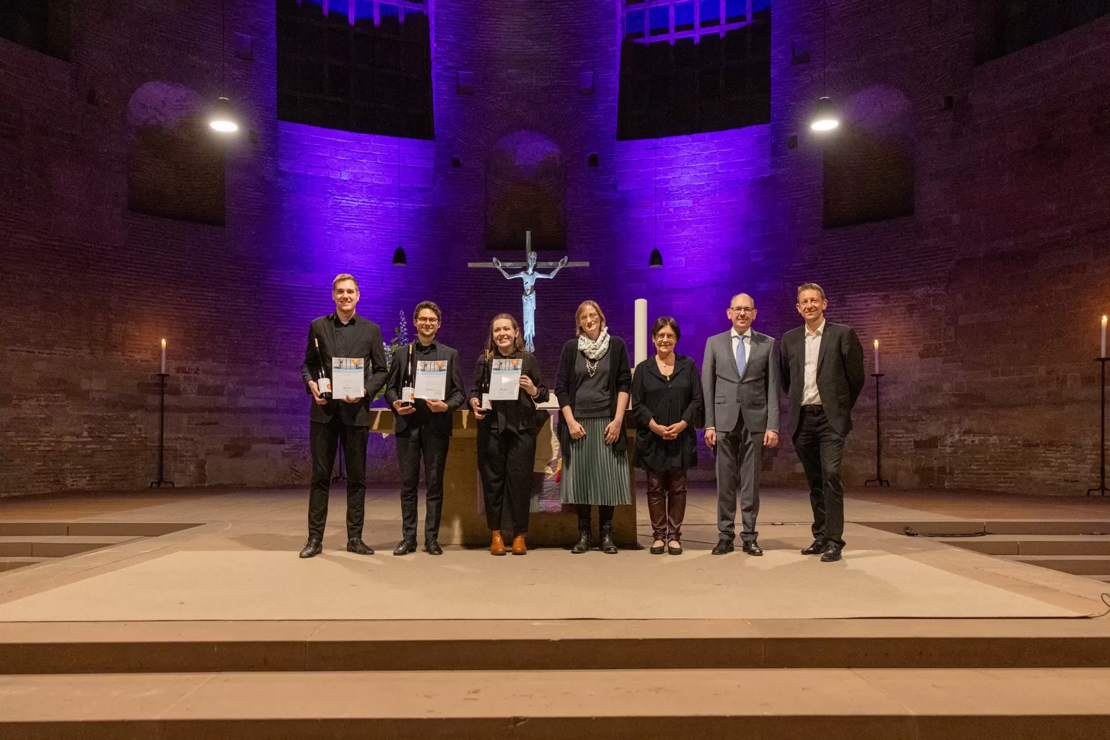 Sieben Personen in formeller Kleidung stehen in einer Reihe in einer Kirche. Die drei Preisträger*innen stehen links und präsentieren ihre Urkunden.
