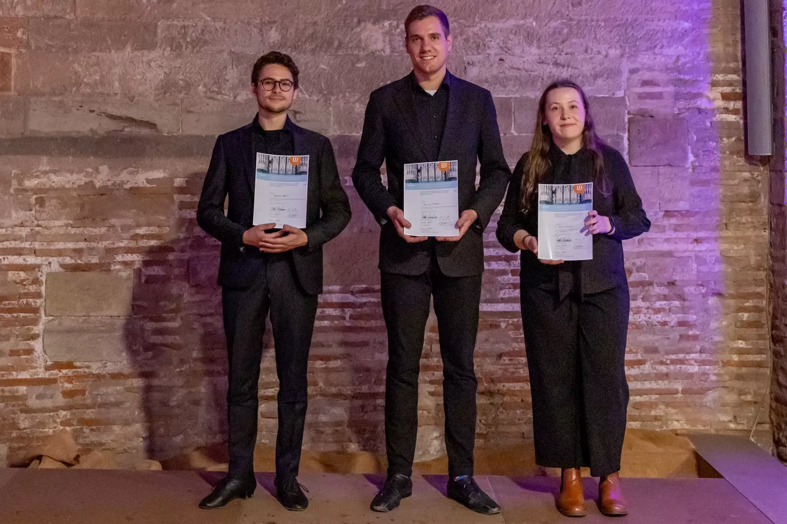Drei schwarz gekleidete stehen nebeneinander und präsentieren ihre Urkunden. Manuel Pschorn steht in der Mitte.