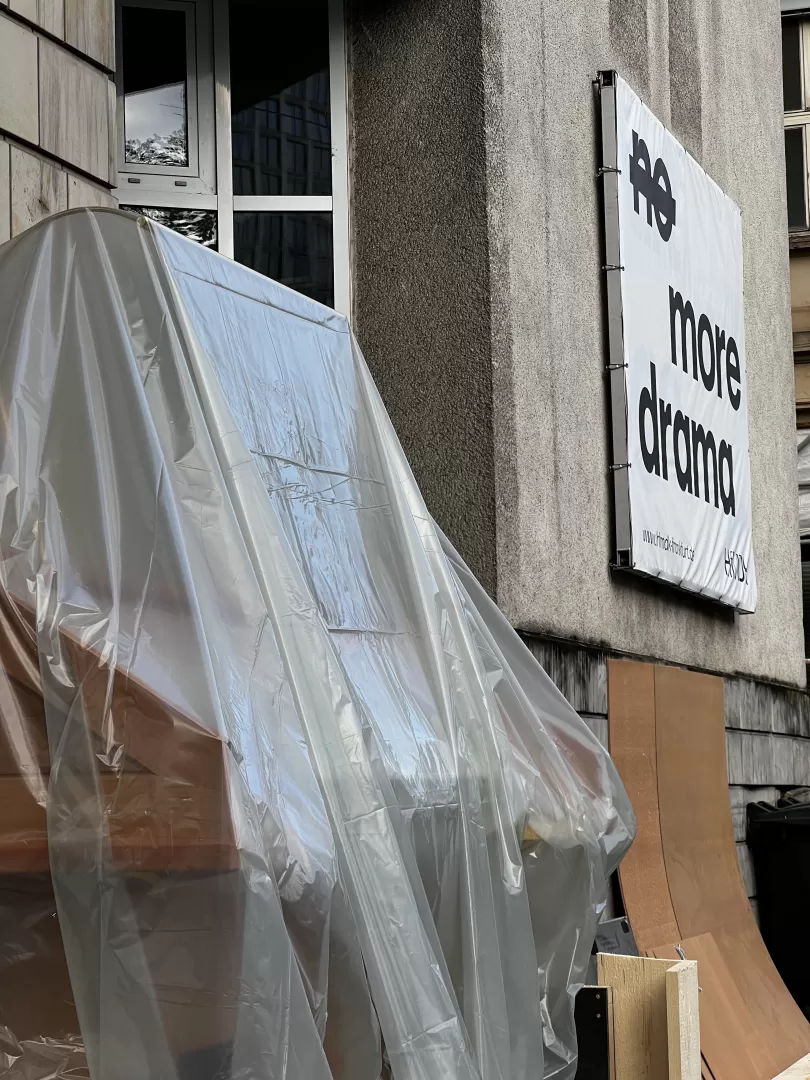 Der unter einer Plane versteckte Spieltisch steht vor dem Hauptgebäude der HfMDK. Im Hintergrund sieht man das Fassadenbanner mit der Aufschrift: "no more drama", dabei ist das Wort "no" durchgestrichen.