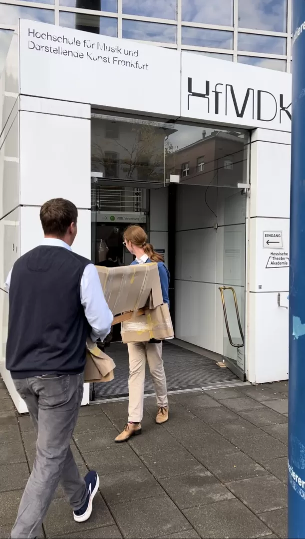 Zwei Personen tragen ein längliches Paket durch den Haupteingang der Hochschule.