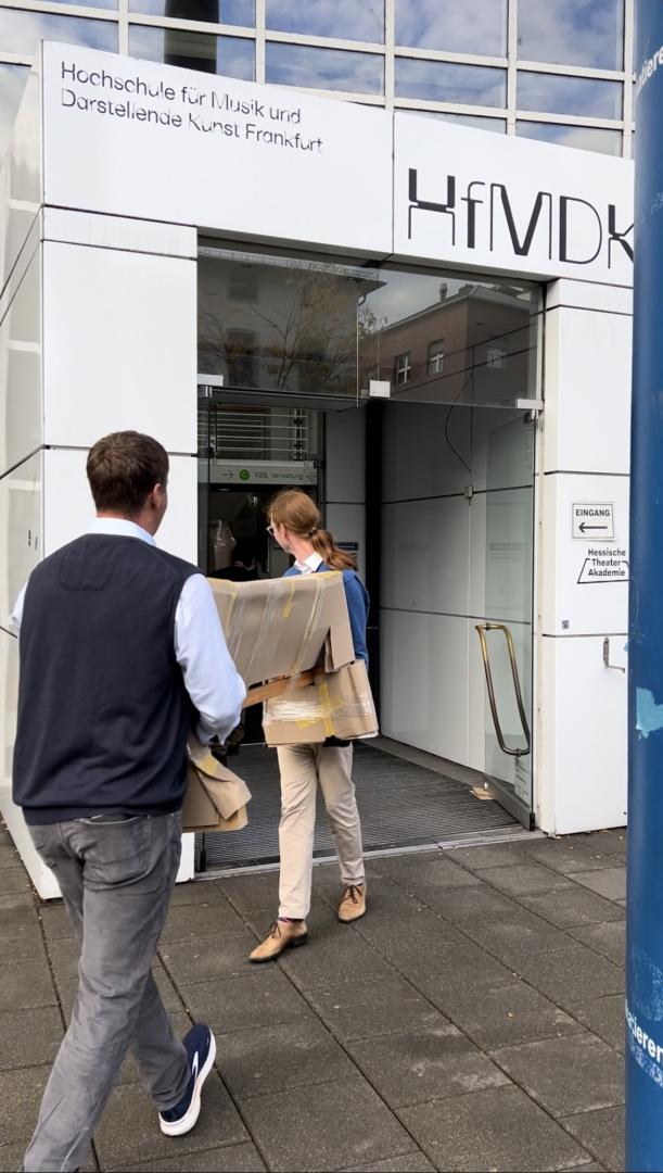 Zwei Personen tragen ein längliches Paket durch den Haupteingang der Hochschule.