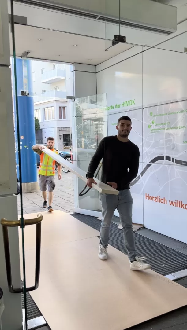 Zwei Personen tragen ein langes Brett durch den Haupteingang.