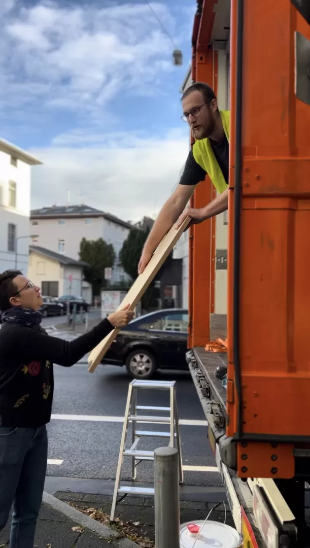 Ein Handwerker gibt einer Studentin eine Holzplatte aus dem LWK heraus an.