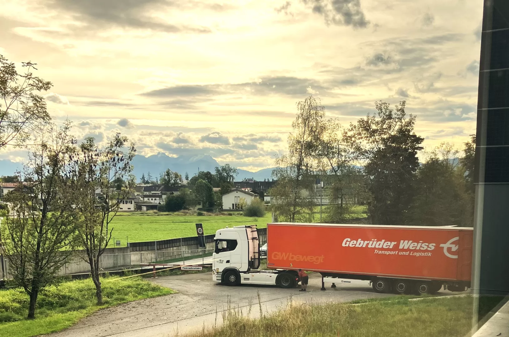 Der Lastwagen steht vor der Werkstatt. Dahinter sieht man den Ort und Berge in der Ferne.