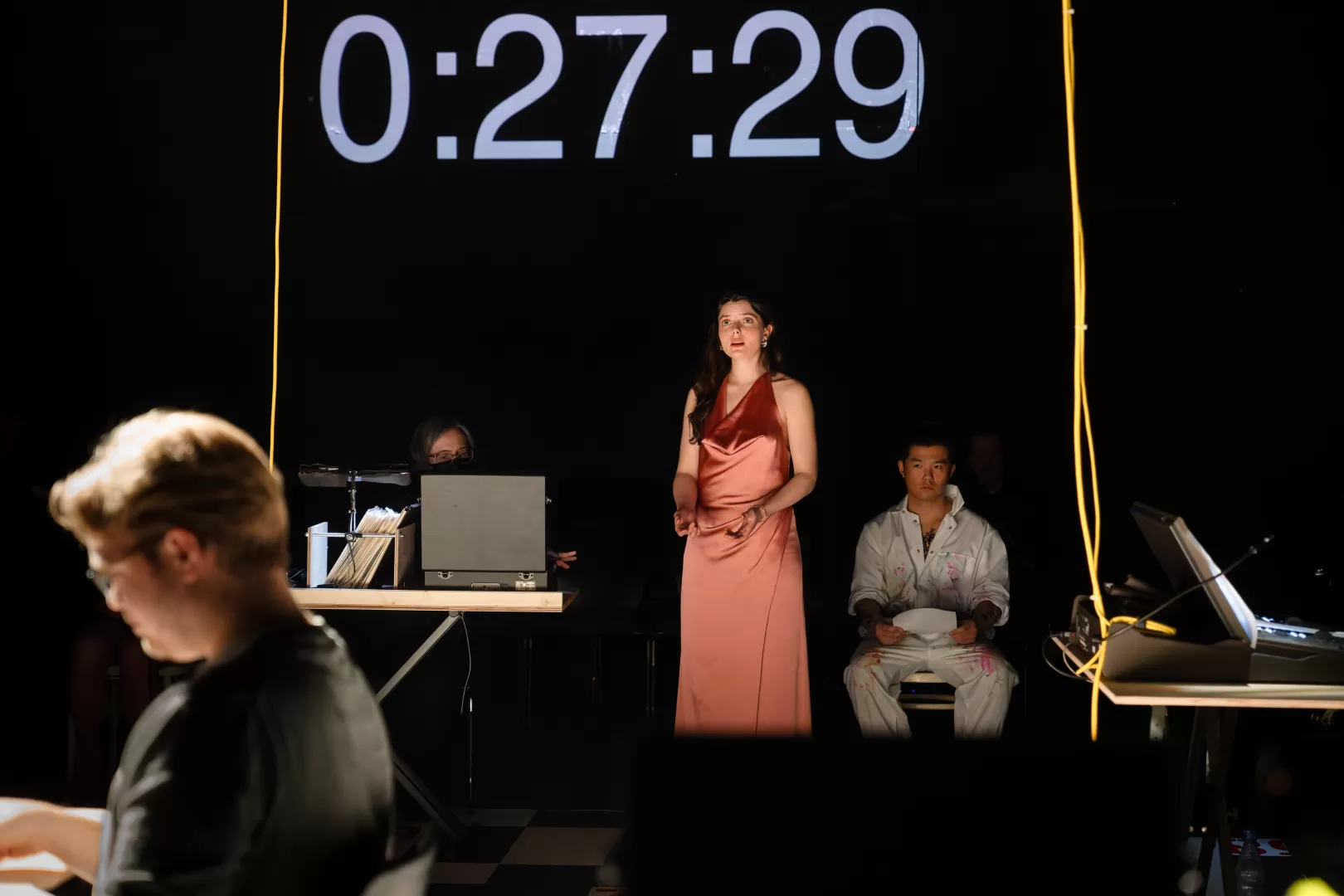 Fotos von Europeras: Eine Sängerin in einem langen Seidenkleid mit traurigem Gesichtsausdruck. Hinter ihr wird die Zahl "0:27:29" groß an die Leinwand projiziert.
