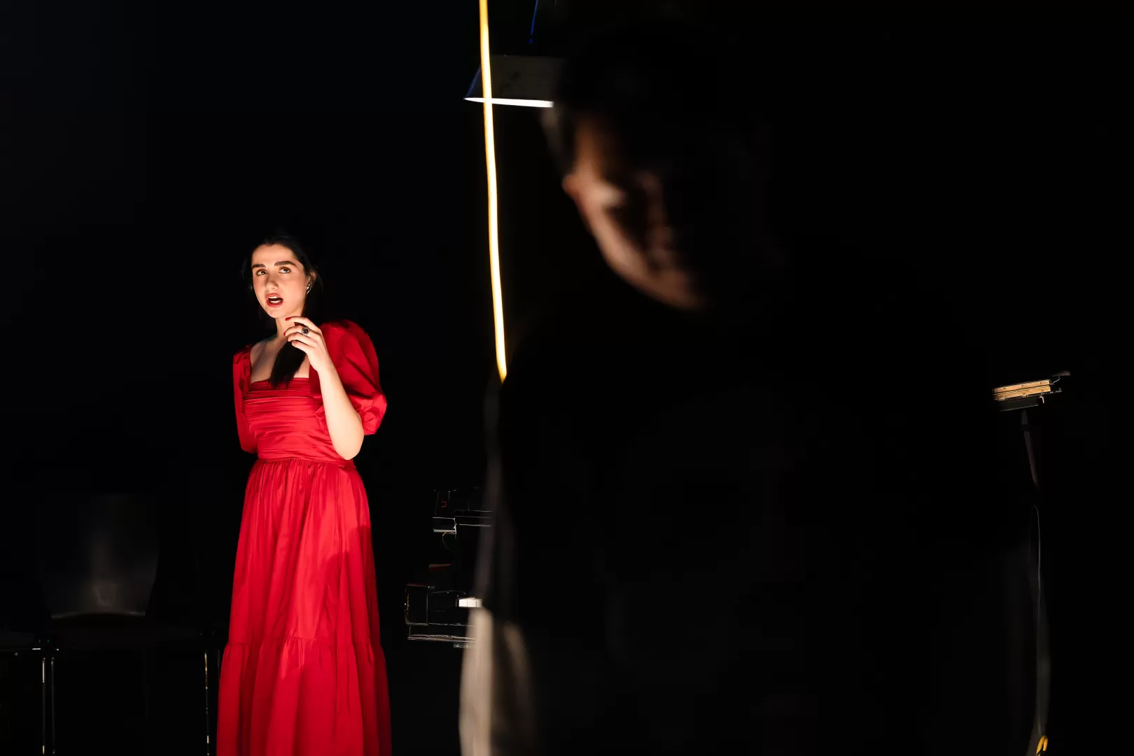 Eine Darstellerin in einem langen roten Kleid. Sie singt und schaut einen anderen Darsteller an, der unscharf im Vordergrund zu sehen ist, mit dem Rücken zu ihr.
