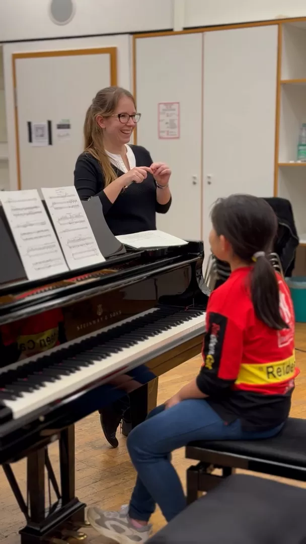Gesa Behrens im Unterricht. Sie steht lächelnd hinter einem Flügel, an dem ein Mädchen sitzt und spricht mit den Anwesenden, die außerhalb des Bildes sitzen.