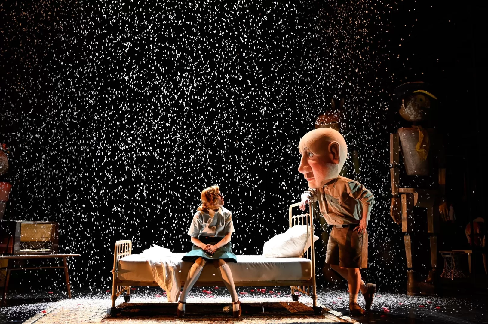 Eine Person sitzt auf einem Bett. Eine andere Person mit Puppenkopf schaut sie an.