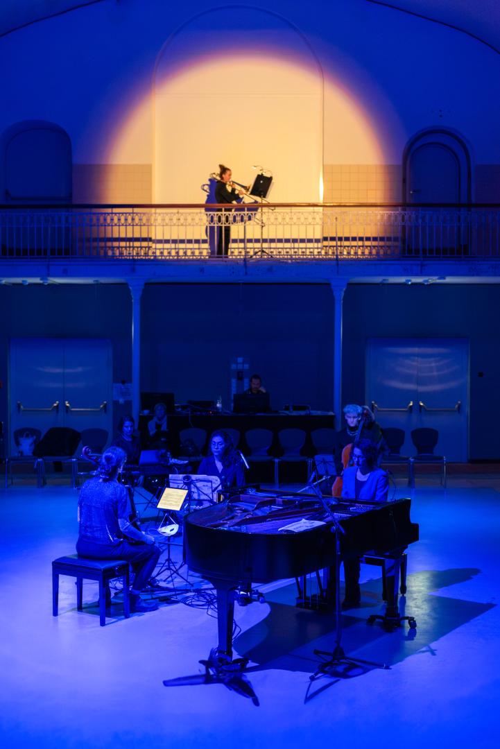 Aufführungsfoto: Die Posaunistin spielt auf einer Empore in einem warmen Spot. Unten spielen die anderen Musiker*innen. Sie sind in blaues Licht gehüllt.