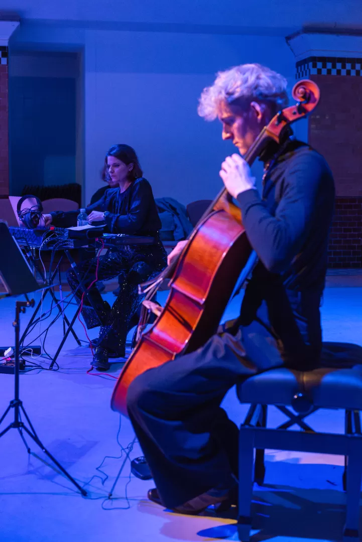 Ein Cellist spielt, im Hintergrund sitzt eine Studentin an der Elektronik. Beide sind umgeben von blauem Licht.