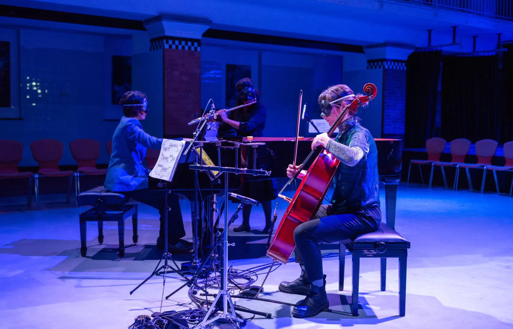 Eine Flötistin, eine Pianistin und eine Cellistin musizieren im Alten Halltenbad. Vor den Augen tragen sie schwarze Masken.