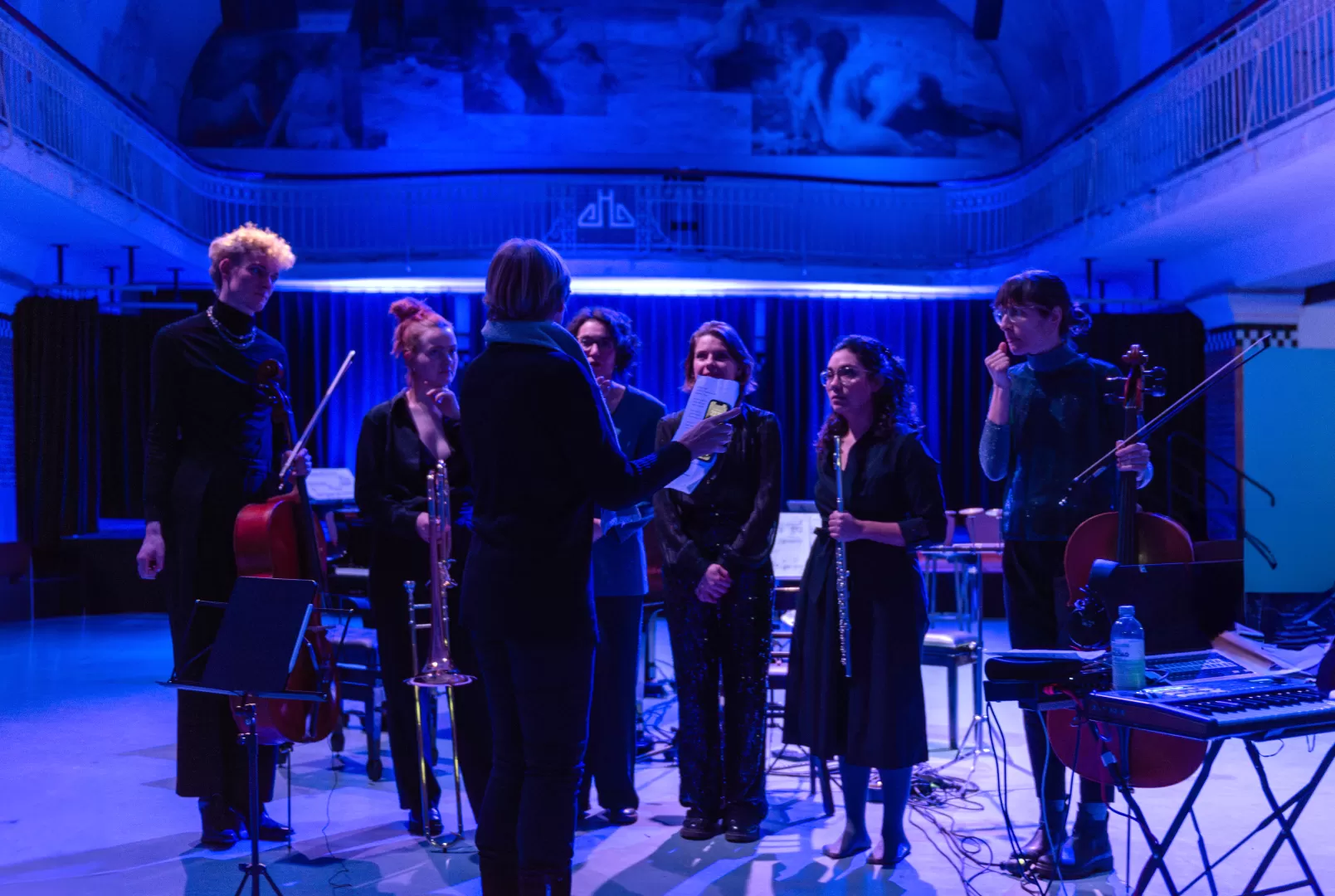 Mehrere schwarz gekleidete Personen stehen in einem Kreis, die Studierenden hören einer Frau aufmerksam zu. Um sie herum ist der Raum in blaues Licht gehüllt.