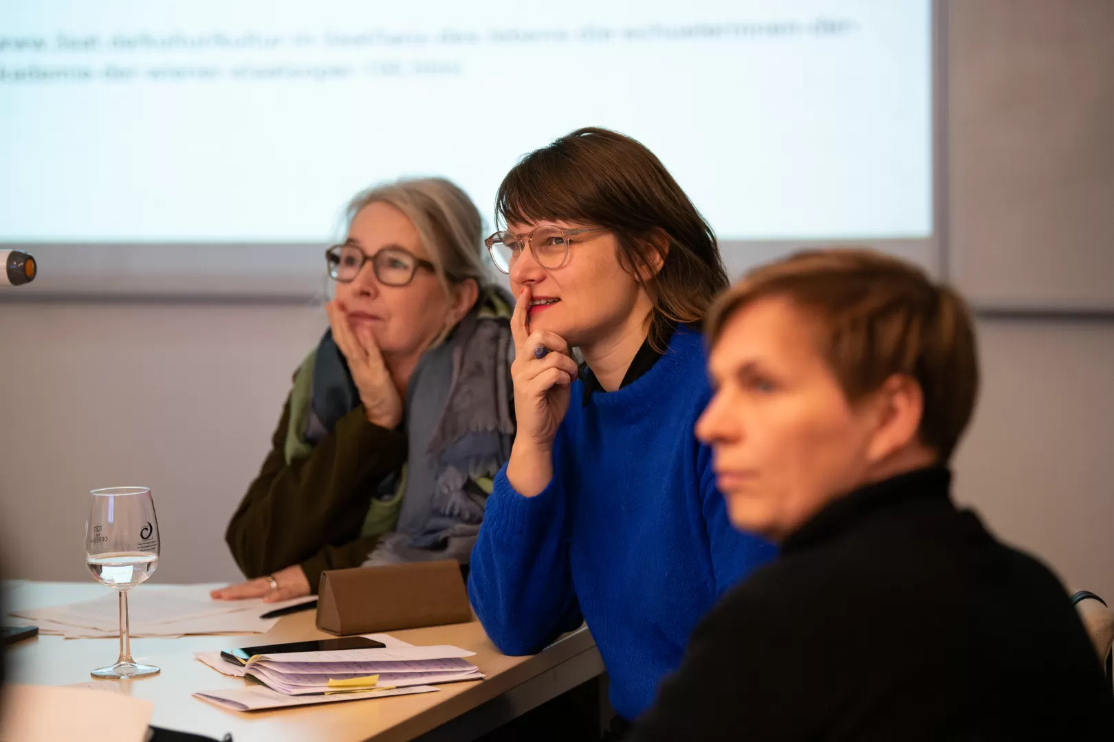 Drei Frauen in einem Konferenz-Setting an einem Tisch, sie blicken gemeinsam nach links, hinter ihnen ist eine Projektion an der Wand zu erkennen.