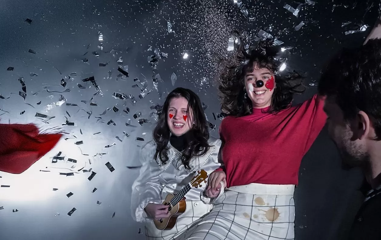 Zwei Frauen sind im Halbporträt zu sehen. Die linke hat lange schwarze Haare, sie trägt eine Satinbluse. Auf ihren Wangen ist jeweils rechts und links ein rotes Herz aufgemalt. In der rechten Hand hält sie eine kleine Gitarre. Die rechte Frau hat ebenfalls langes dunkles Haar, sie trägt einen roten Pulli und hat eine schwarze Clownsnase auf der Nase und rote und weiße Farbe auf den Wangen. Beide Frauen lachen. Links von ihnen und vor ihnen ist silberner Flitter zu sehen.