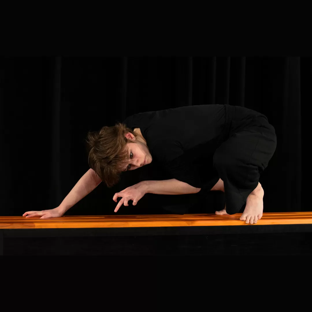 Aus „Tiere und Etüden“, schwarze Kleidung, auf Händen und Füßen auf einer Bank stehend, im Hintergrund ein schwarzer Vorhang