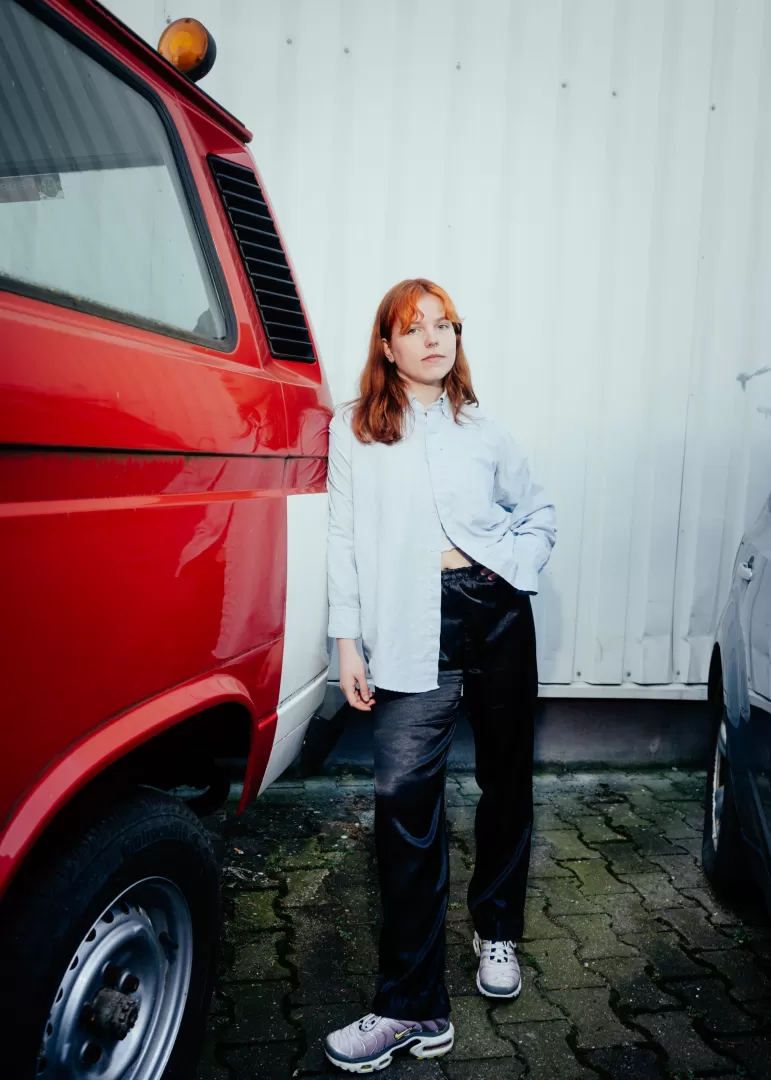 junge Frau mit roten Haaren. Sie trägt ein Hemd und lehnt sich an ein rotes Auto