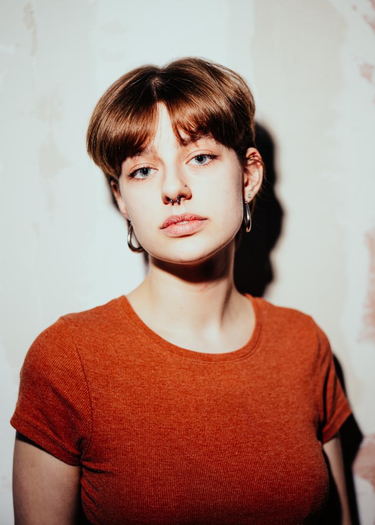 Portrait Paula Marie Schulze, orangefarbenes T-Shirt, im Hintergrund eine kahle, beige Wand