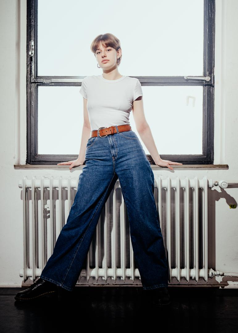 Totale Paula Marie Schulze, blaue Jeans und ein weißes T-Shirt, im Hintergrund ein Fenster und ein weisser Heizkörper 