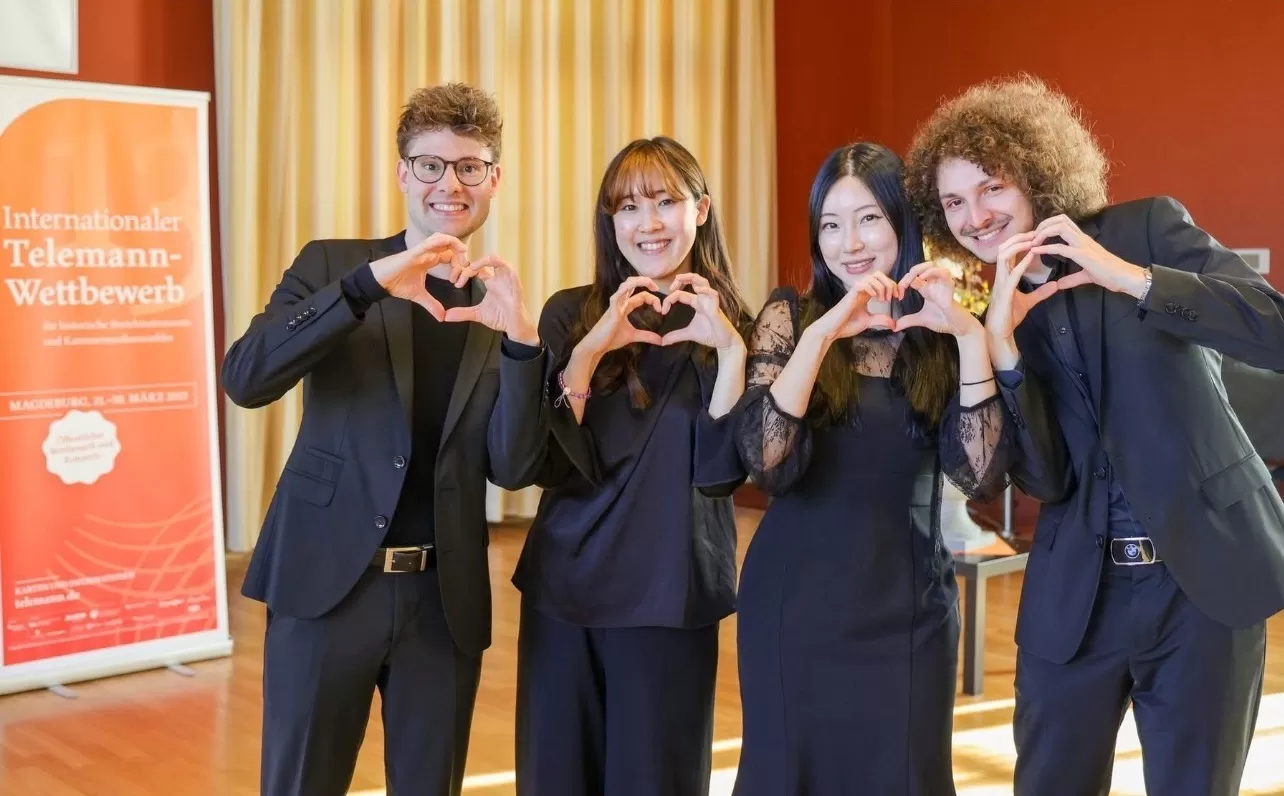 Die vier Musiker*innen bilden Herzen mit ihren Händen und lächeln in die Kamera. Sie stehen vor einem Roll-up des Wettbewerbs und tragen schwarze Kleidung.