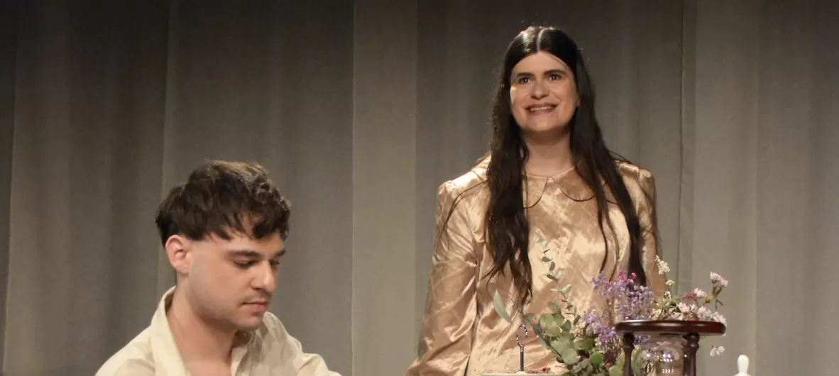 Links im Bild ist der Kopf eines Mannes zu sehen. Er hat kurze braune Haare und blickt nach unten. Rechts von ihm steht eine Frau. Sie ist bis zur Hüfte zu sehen. Sie hat lange braune Haare, lächelt und schaut nach vorn.