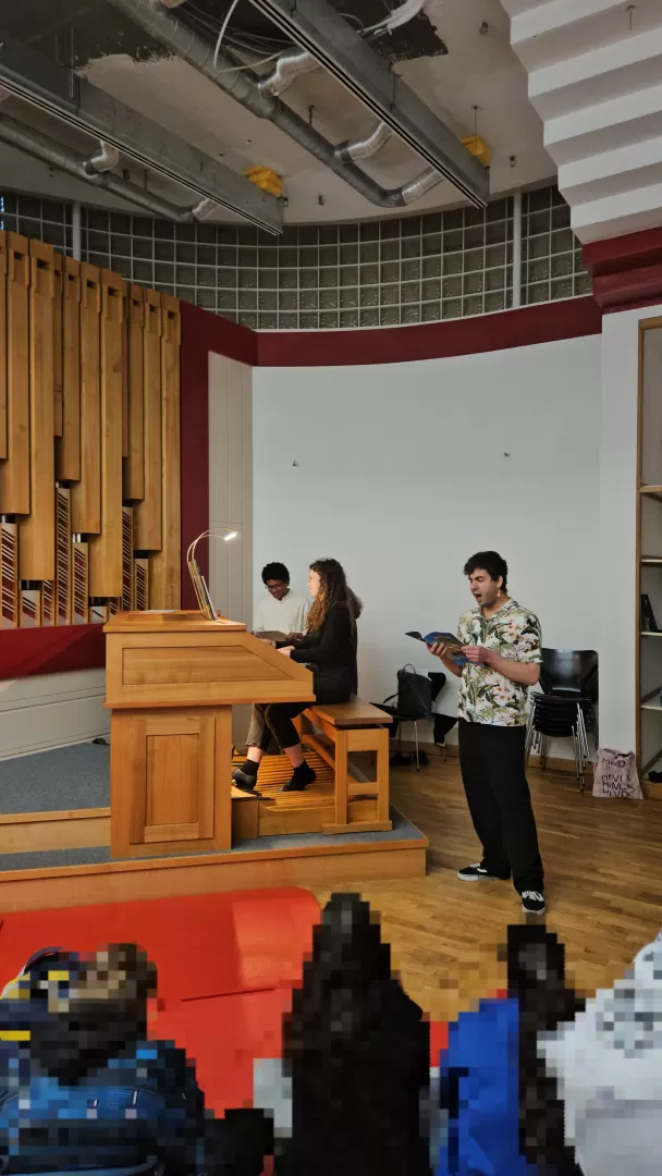 Blick in Orgelraum. Student liest laut energisch den Text, andere Studentin sitzt an der Orgel. Schulklasse hört zu.