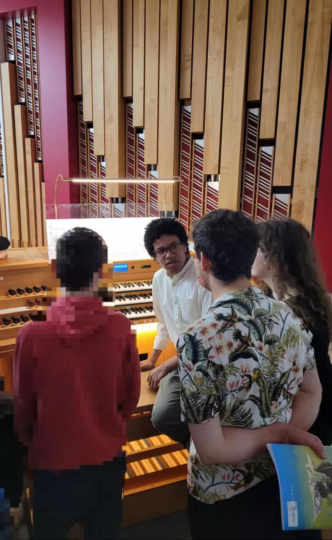 Ein Student sitzt auf der Orgelbank, zwei weitere Studierende stehen daneben und unterhalten sich mit einem Schüler