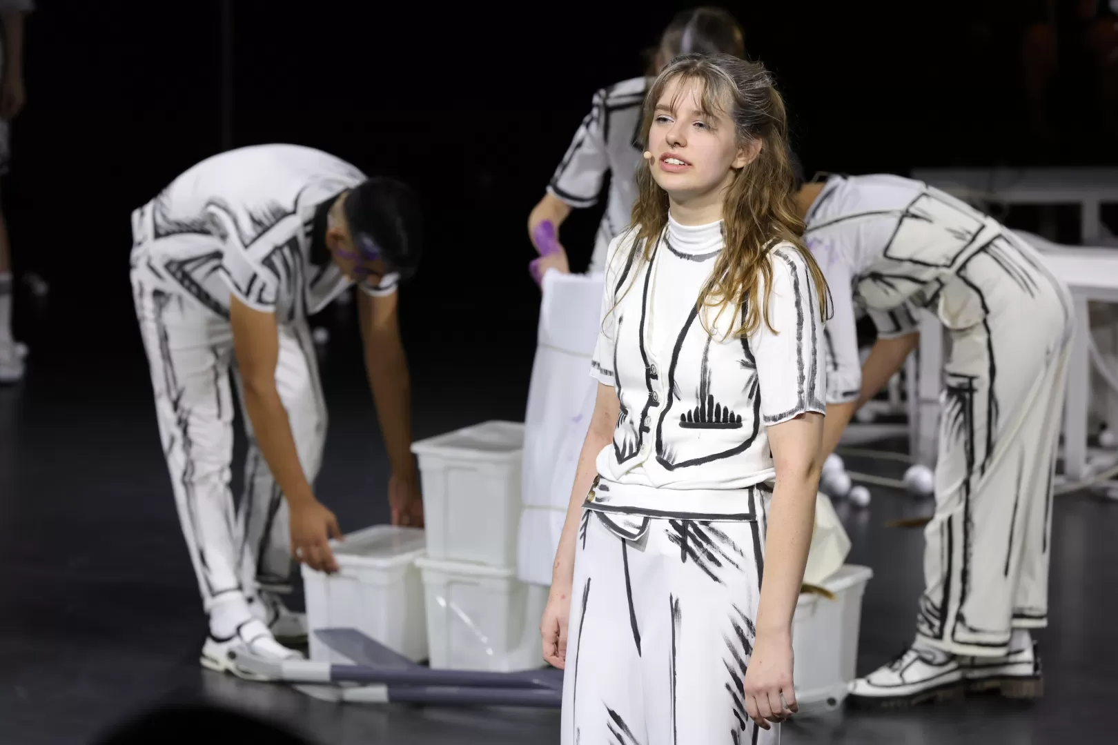 Paula Marie Schulze in einer weißen Weste und Anzugshose mit comicartiger schwarzer Umrandung, im Hintergrund ein Stapel an Plastikboxen und weitere Spielende