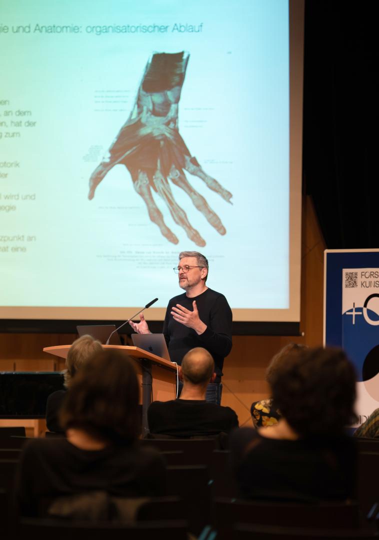Ein Mann trägt im Kleinen Saal der HfMDK am Pult vor, hinter ihm ist ein schematisches Bild von einer Hand auf die Leinwand projiziert.