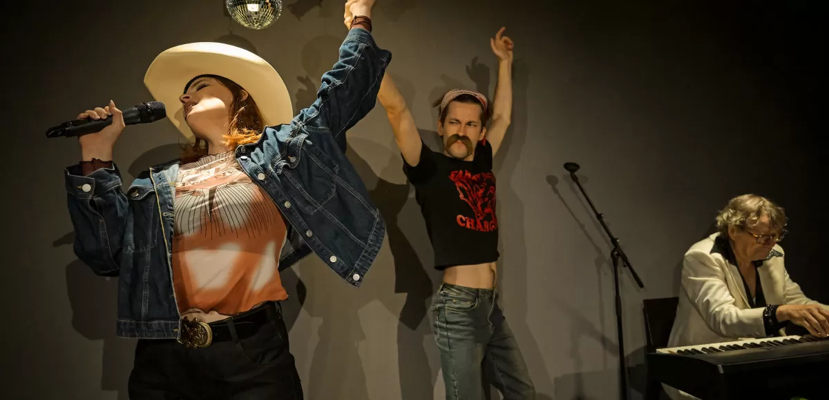 Zwei Leute singen und tanzen dabei auf einer Bühne. Rechts sitzt jemand am Klavier und spielt.