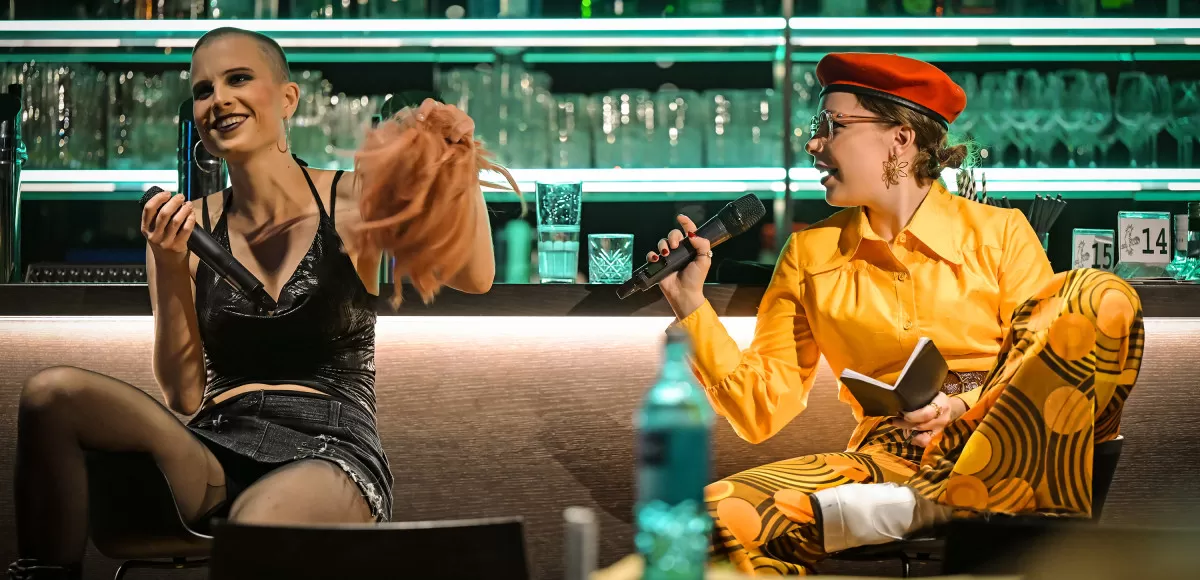 Zwei Leute sitzen vor einer Bar. Die rechte Person hat ein Mikro und schaut zu der linken.