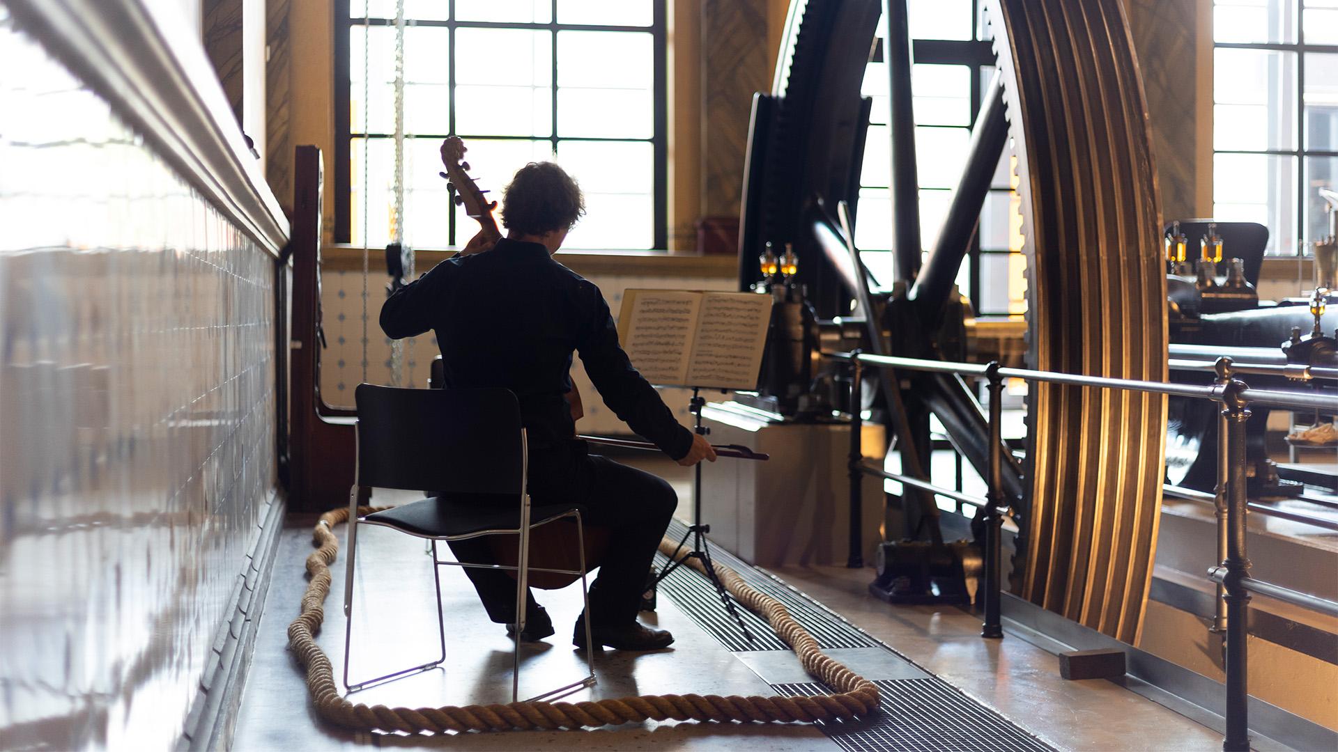 Ein Cellist spielt neben einem großen Zahnrad.