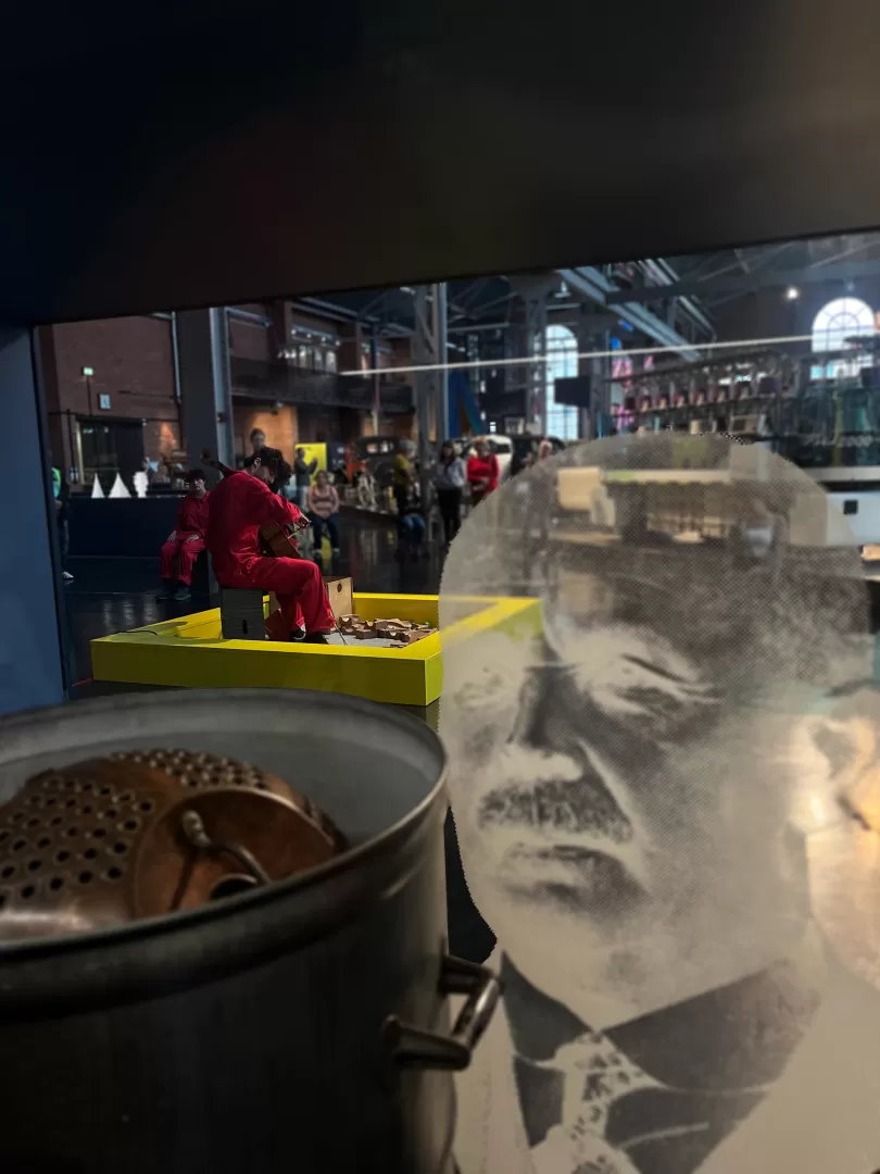 Ein Cellist musiziert im Industriemuseum, während mehrere Besucher zusehen, durch eine Vitrine hindurch fotografiert.
