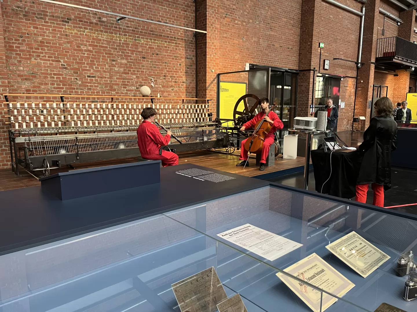 Rot-schwarz gekleidete Menschen musizieren im Industriemuseum. Zwei Personen vor einer Backsteinwand spielen Oboe und Cello. Davor steht eine Person am Laptop.