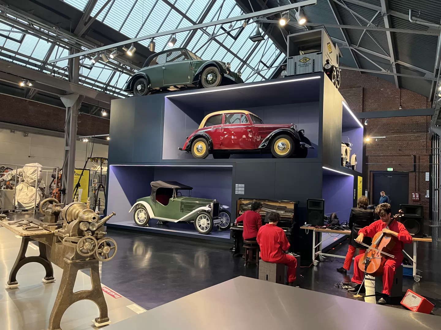 Mehrere Menschen in roten Overalls musizieren mitten im Industriemuseum. Hinter ihnen sind alte Autos ausgestellt. Im Vordergrund sieht man eine Werkbank.