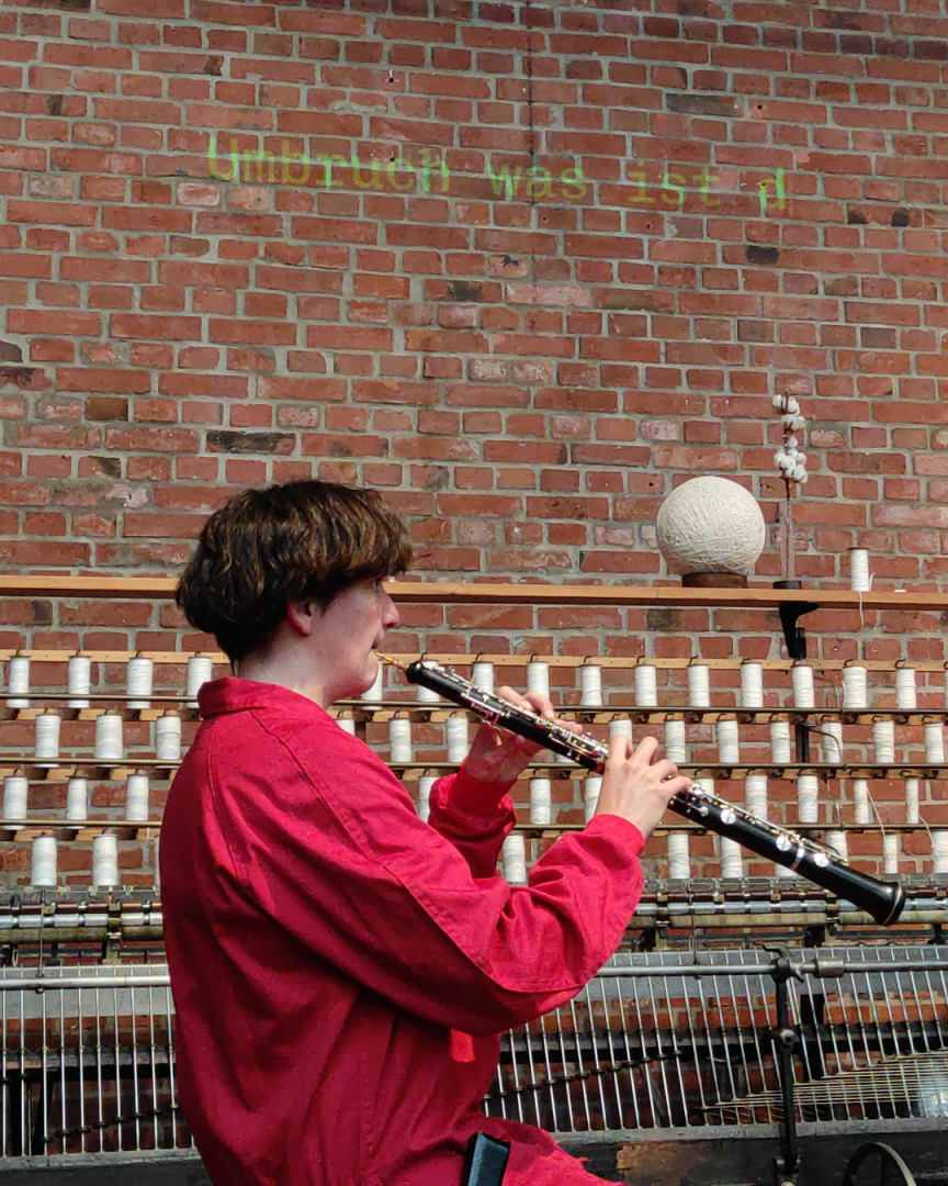 Eine Person in einem roten Overall spielt Oboe. Auf die Backsteinwand hinter ihr wird der Text projiziert: "Umbruch was ist d..."