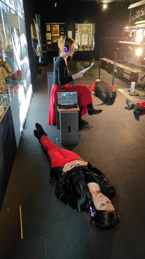 Eine Person sitzt inmitten einer Ausstellung und liest etwas vor. Weitere Personen liegen auf dem Boden und tragen Kopfhörer.