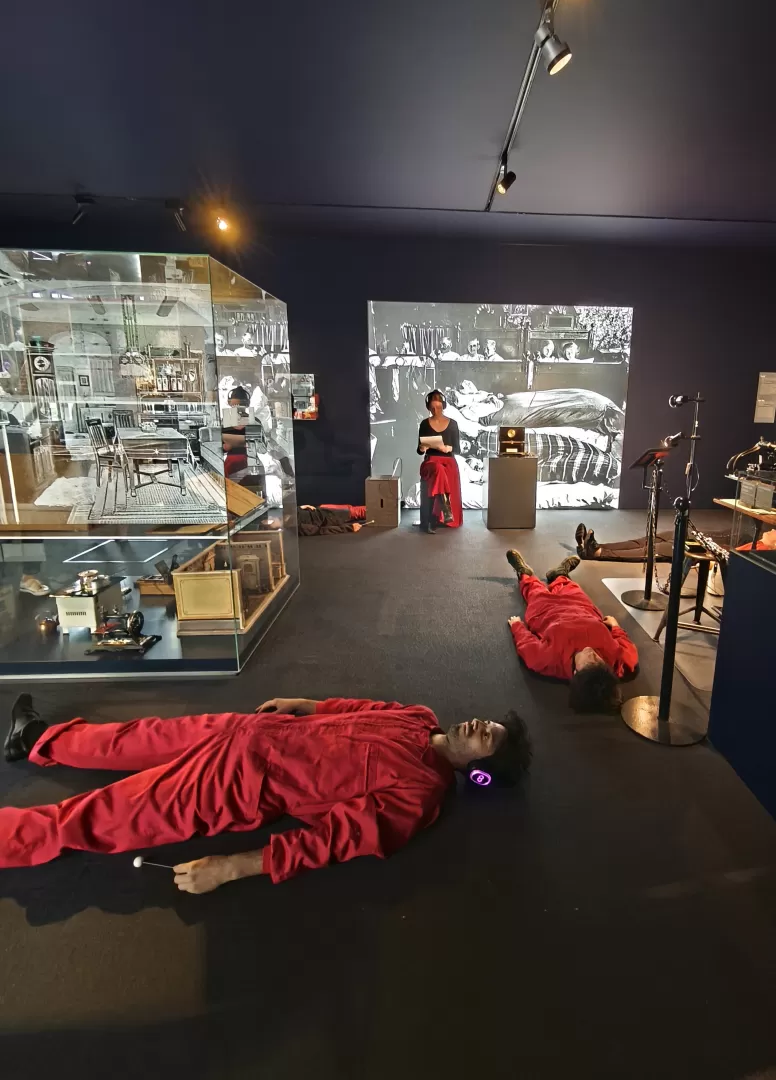Mehrere Menschen in roten Overalls liegen mit Kopfhörern auf dem Boden in einer Ausstellung. Eine Person sitzt auf einem Klotz und liest etwas von einem Blatt Papier ab.