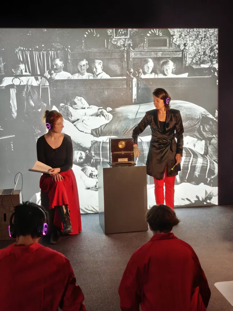 Rot-schwarz gekleidete Menschen mit Kopfhörern im Industriemuseum. Eine Person berührt eine antik aussehende Uhr.