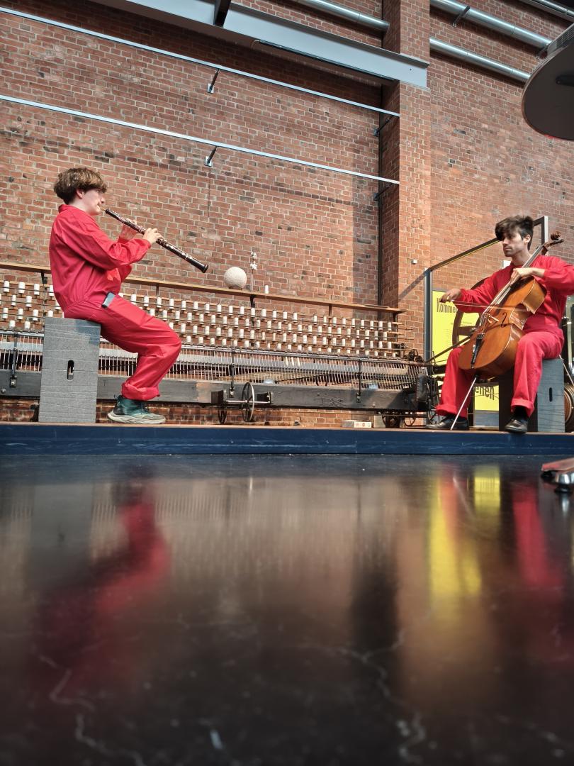 Zwei Studierende in roten Overalls sitzen vor einer Backsteinwand im Museum und spielen Oboe bzw. Cello.