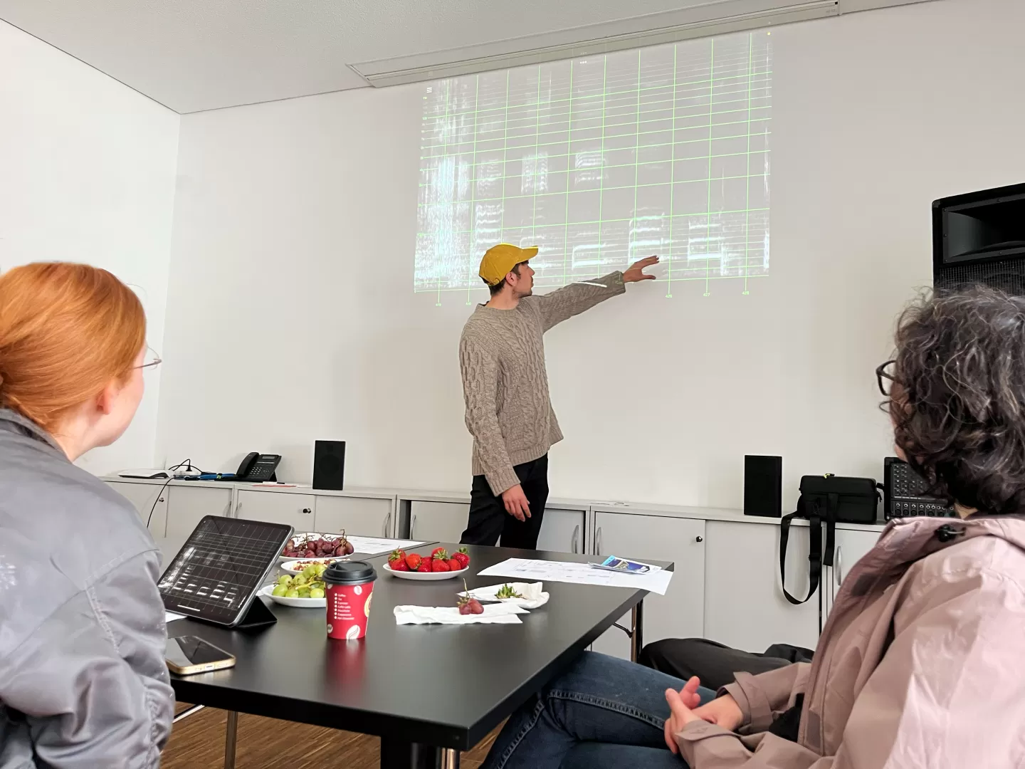 Spektogramme werden an eine weiße Wann gebeamt. Ein Mann steht davor und zeigt darauf. Vorne sind andere Menschen zu sehen, die dort sitzen und zuhören.