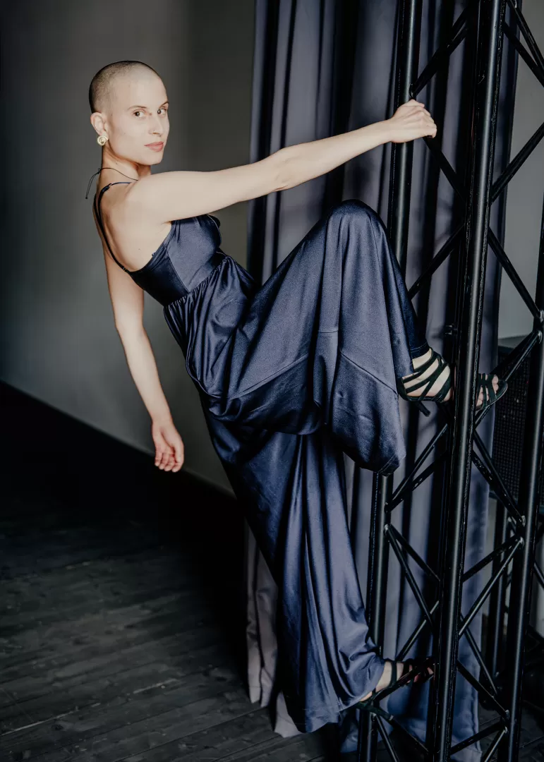 Lisa Freiberger steh mit einem Fuß auf einer Säule und hält sich mit der Hand dort fest. Ihr Körper ist eingedreht, der Blickt geht zur Kamera. Sie trägt einen blauen Jumpsuit.