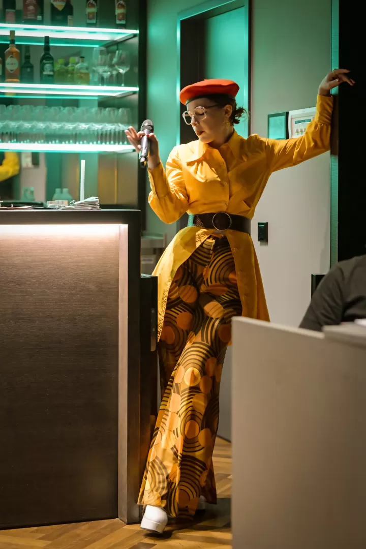 Frau in gelber Kleidung mit Mikrofon an einer Bar.