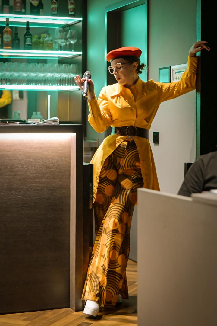 Frau in gelber Kleidung mit Mikrofon an einer Bar.