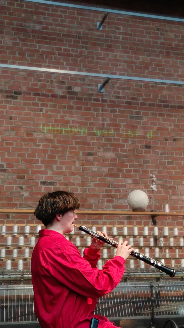 Eine Person in einem roten Overall spielt Oboe vor einer Backsteinwand