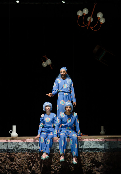 Zwei Menschen sitzen auf einer Kante vor einem dritten. Alle schauen nach rechts und tragen blaue Perücken und blaue Uniformen