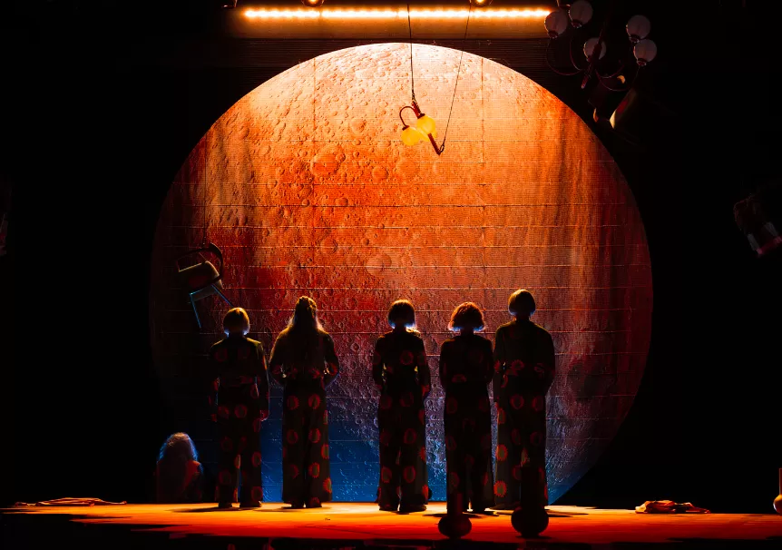 Fünf Personen stehen im Schatten. Hinter ihnen ist ein orangen angeleuchteter Mond